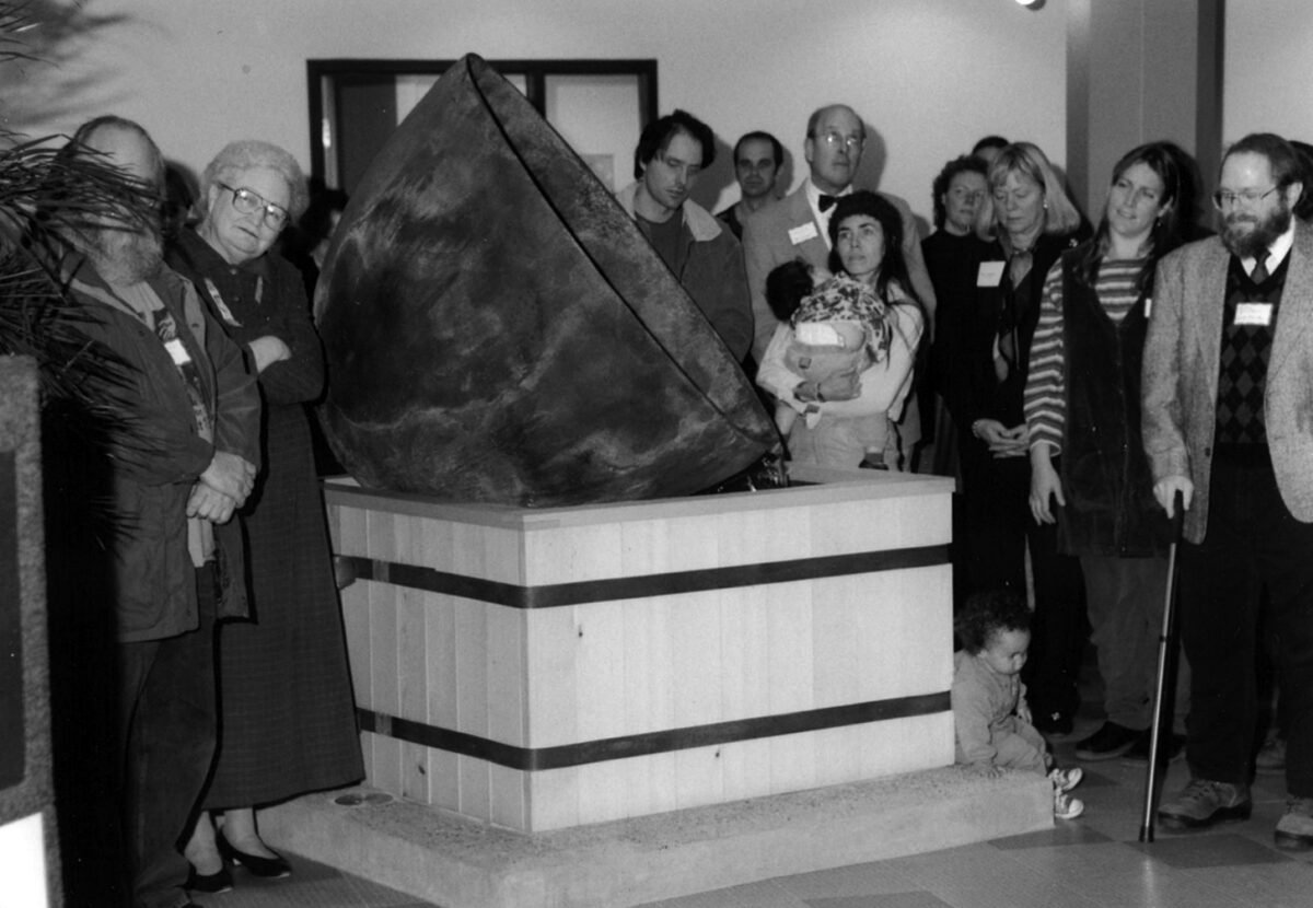 Group of people gathered around a large, wedge-shaped stone sculpture on a pedestal in a gallery or museum lobby, with onlookers watching intently.