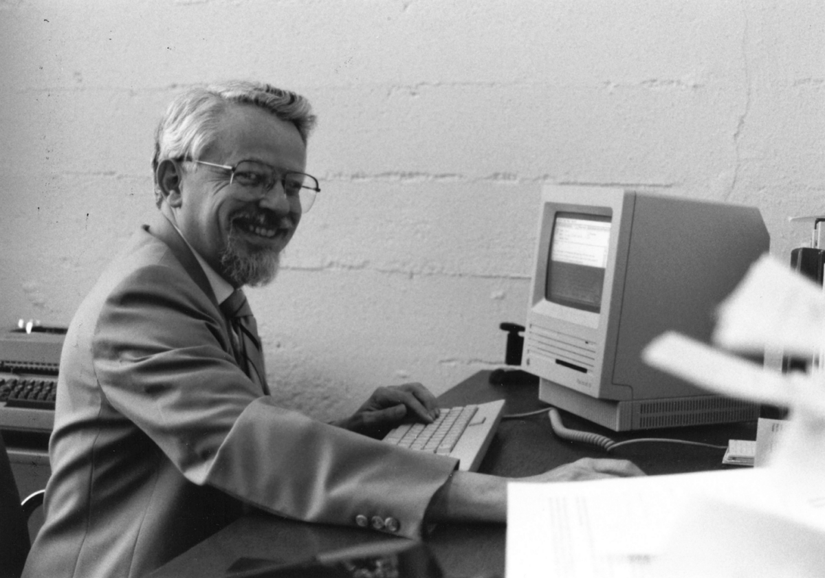 Man with glasses and a beard sits at a desk, typing on a vintage computer and smiling at the camera.