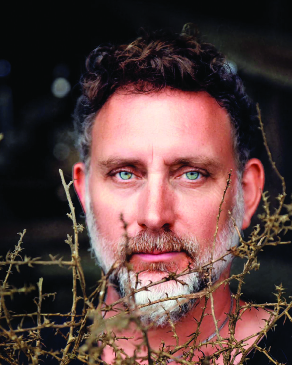 Close-up of a middle-aged man with blue-green eyes and gray stubble, framed by thorny branches.