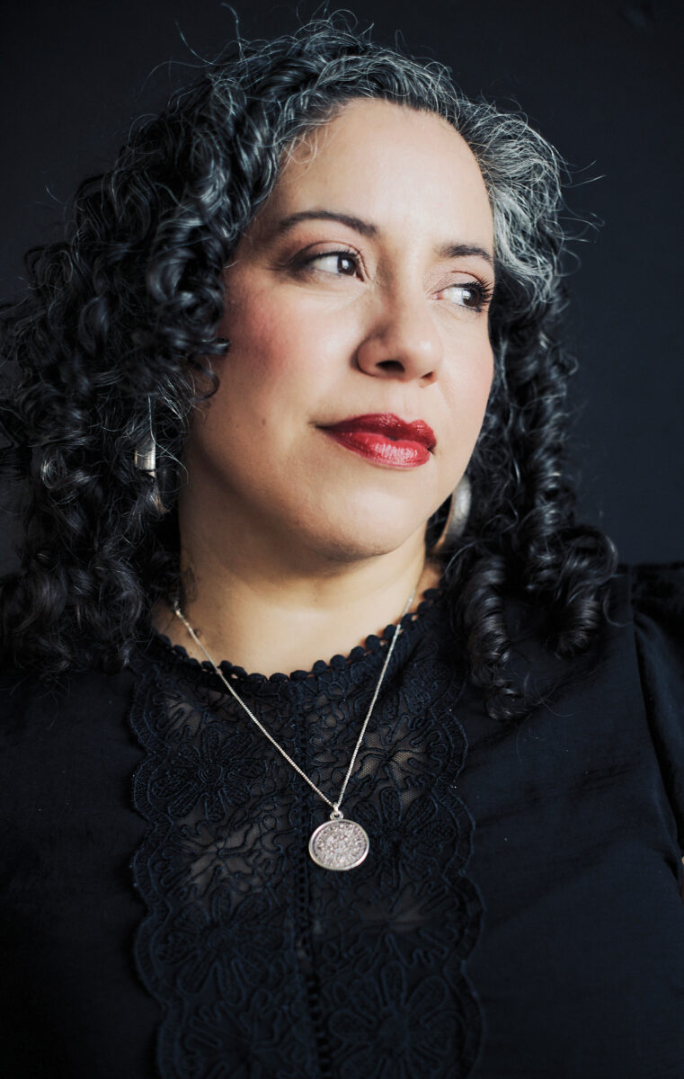 Close-up portrait of a woman with dark curly hair, wearing a black lace top and a silver pendant, looking off to the right.