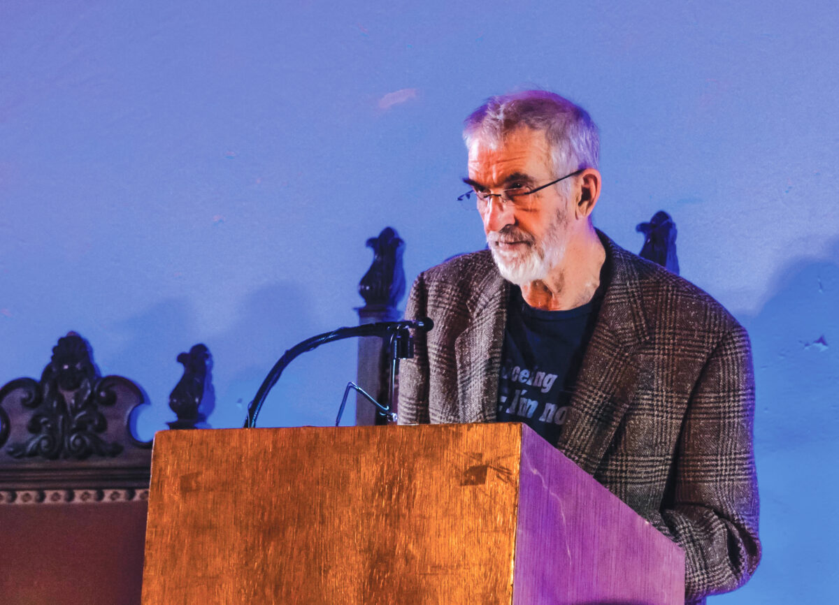 Older man with glasses and a gray beard speaking at a wooden podium with a microphone.