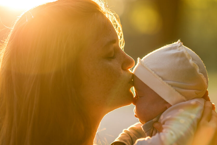 A mother kisses her bundled infant outdoors in warm evening light