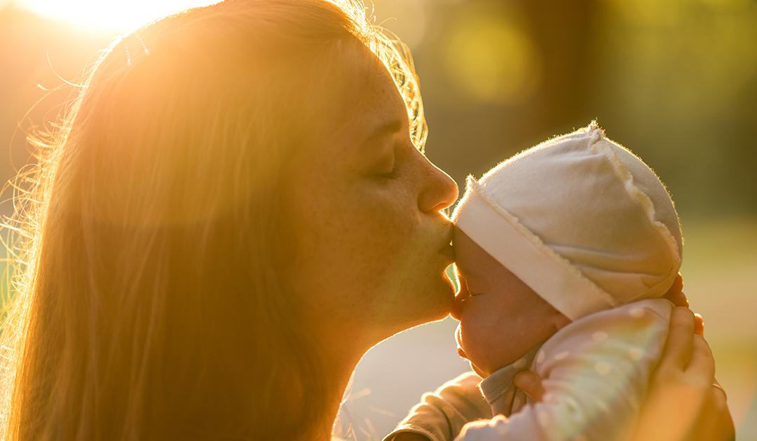 A mother kisses her bundled infant outdoors in warm evening light
