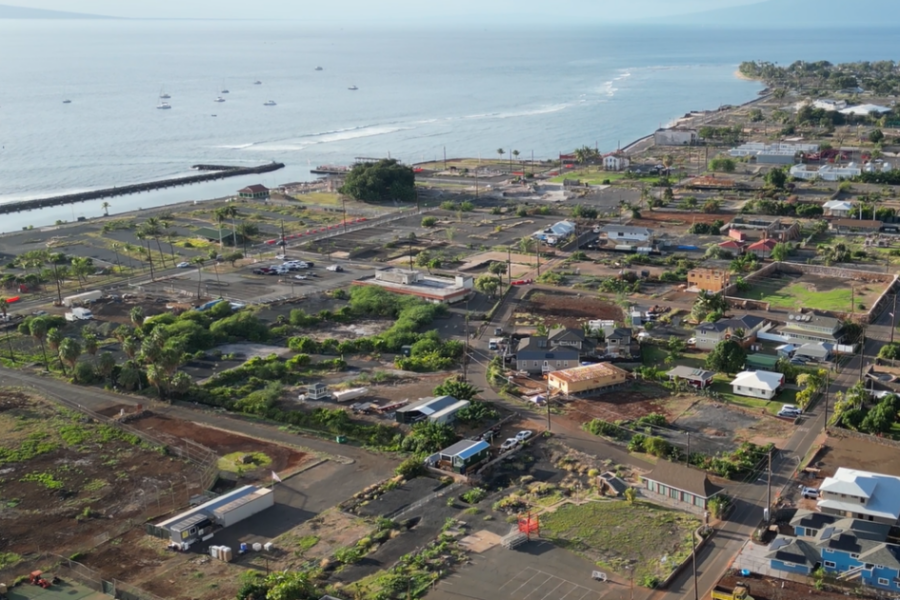 Aerial view of Lāhainā, Maui, showing homes, roads, open lots, and areas of rebuilding along the coastline, with the Pacific Ocean in the background.