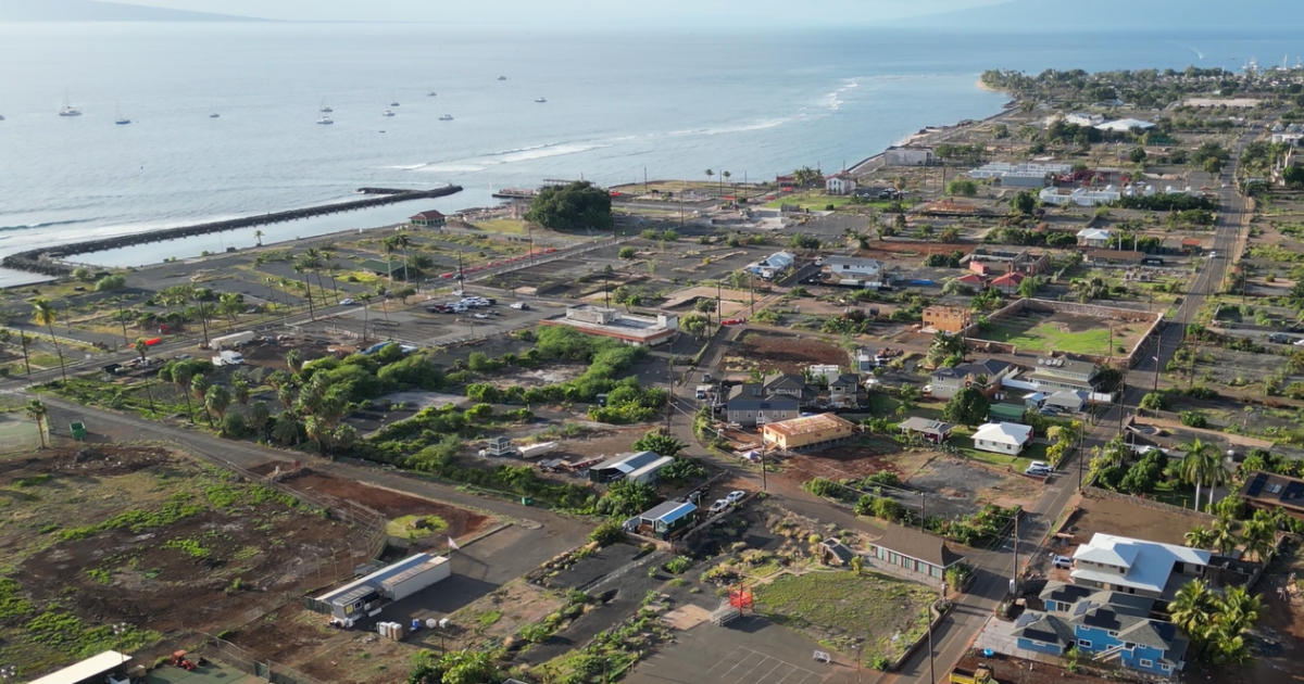 Aerial view of Lāhainā, Maui, showing homes, roads, open lots, and areas of rebuilding along the coastline, with the Pacific Ocean in the background.