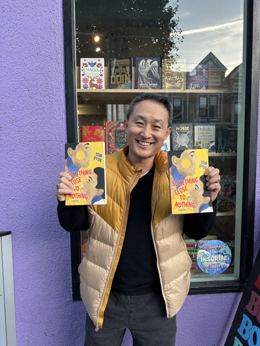 A photo of Tom Pyun who is smiling and wearing a vest, holding two copies of the book Something Close to Nothing and standing in front of a bookstore window.