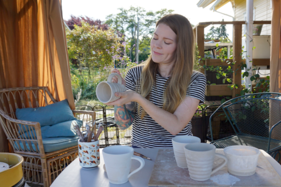 Jessica Mudd holds a handmade ceramic mug while standing beside a table of pottery and art supplies in a sunny outdoor studio space.