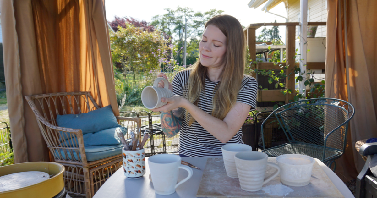 Jessica Mudd holds a handmade ceramic mug while standing beside a table of pottery and art supplies in a sunny outdoor studio space.