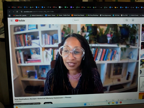 Stephanie Helms Pickett with long dark hair and glasses speaking during a virtual presentation, with bookshelves and plants in the background.