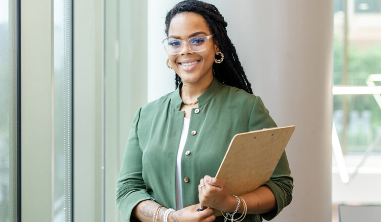 Smiling woman wearing glasses holds a clipboard in a bright office