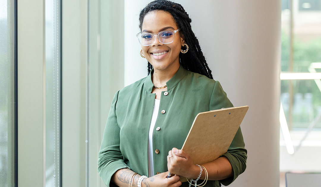 Smiling woman wearing glasses holds a clipboard in a bright office