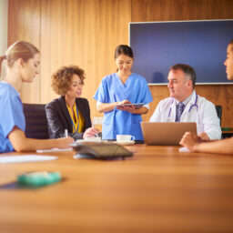 Healthcare professionals meeting around a conference table, with a doctor using a laptop and nurses reviewing notes