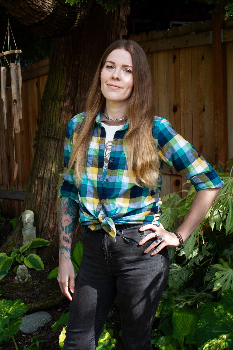 Jessica Mudd stands in a garden wearing a colorful plaid shirt, with trees, greenery, and a wooden fence behind her.