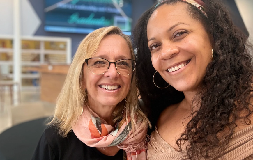 Alicia Betha (right) and Diane Richard-Allerdyce (left) smile together for a photo indoors. Betha wears a beige top and patterned headband, while Richard-Allerdyce wears glasses and a colorful scarf. A softly blurred background with blue tones frames the photo.