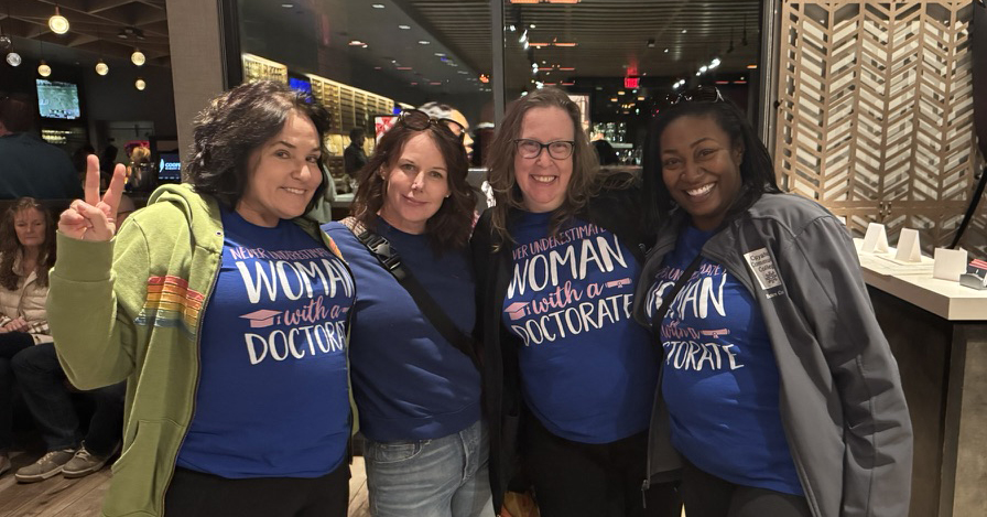 A banner image showing four women posing in a restaurant. Each is wearing a shirt that says "Never Underestimate a Woman With a Doctorate"