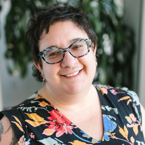 Amy Lesen- Smiling person with short dark hair and glasses wearing a floral-patterned shirt