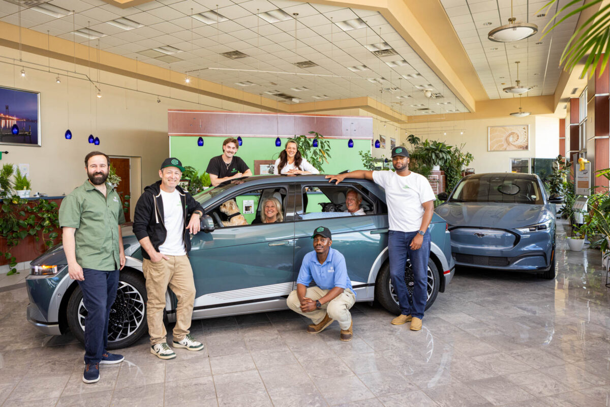 Six people pose around an electric car in a showroom, with another electric car and a dog visible inside